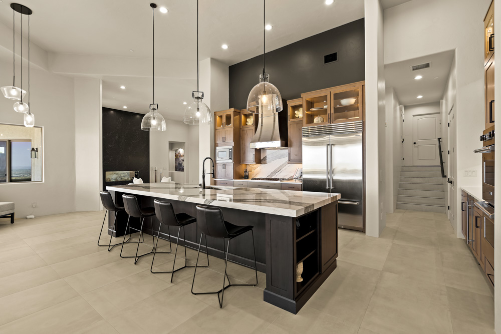 A spacious kitchen featuring a large dark island with a marble countertop, black bar stools, and pendant lighting. The background includes a stainless steel refrigerator, range hood, and wooden cabinets, with a hallway leading to upper levels visible in the distance.