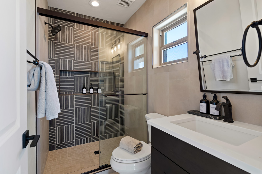 A modern bathroom featuring a walk-in shower with dark wood-look tile walls, a glass sliding door, and a built-in shelf. The space includes a white toilet, a double sink vanity with a black cabinet, and a large mirror with black hardware.